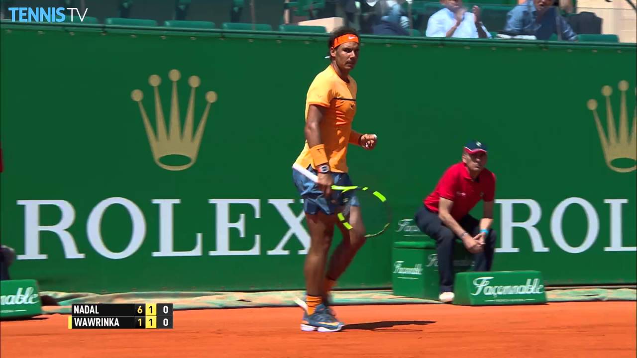 Foto do tenista Rafael Nadal em uma partida, ele está segurando uma raquete de tênis, está andando, por isso está um pouco borrada a imagem. Ao fundo dele tem um painel verde da Rolex.