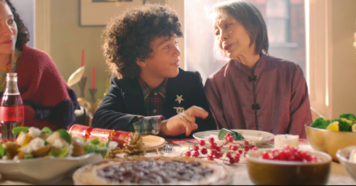 Essa imagem é de um comercial, em que há um garoto sentado a mesa posta, com duas senhoras ao seu lado e no canto esquerdo tem uma garrafa da Coca-Cola.
