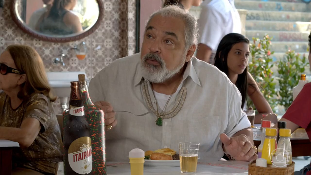 A foto é de uma cena em que o personagem está em um bar, comendo um lanche e bebendo cerveja, na mesa está uma garrafa de Itaipava.